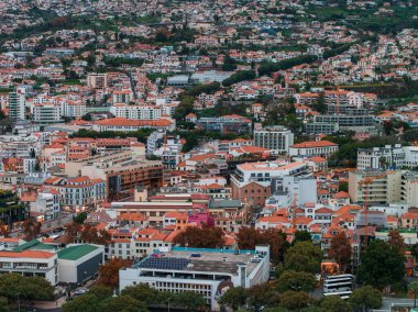 Teraslı Funchal yamaçlarında kırmızı kil çatılar, orta yükselen oteller, eski duvarlar ve güneş panelleri ve bir teleferik istasyonu olan bulutlu ışık altında ağaçlarla kaplı caddeler yer alıyor.