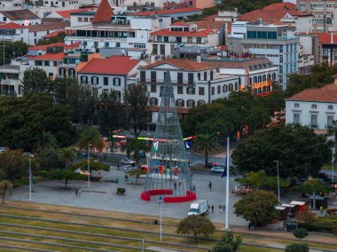Funchal, Madeira 'da bir meydanın manzarası yükselmiş, akşam ışığı. Kırmızı bir üsteki uzun bir konik metal ağaç palmiye ağaçları ve gezinti güvertesinin yanında durur..