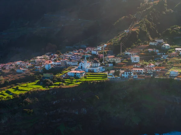 Beyazlatılmış evler Madeira, Portekiz 'deki bir kilisenin etrafında toplanıyor. Teraslar bir vadiye ve Atlantik kıyısına çıkar. Orman dağları ve öğleden sonra ışığı vardır..