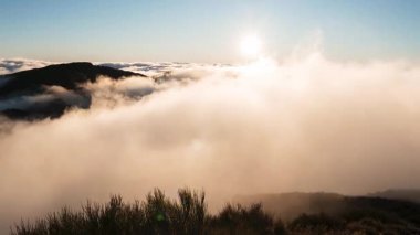 Hava aracı Madeira 'da güneş doğarken bulutların üstündeki dağ sırtlarını gösteriyor. Pico do Arieiro yakınlarındaki tepelerin üzerinden sis yuvarlanıyor, kayalık çalılar ön planda ve sıcak altın ışıkla.