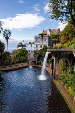 Portekiz, Madeira, Funchal 'daki Jardim Monte Sarayı' nda kemerli bir geçidin altında şelale yapılmış bir adamla kıvrımlı bir gölet. Monte Sarayı Atlantik görünür bir şekilde yukarıda duruyor..