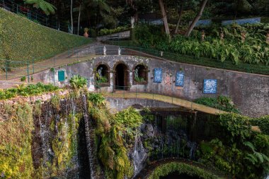 Teraslı bahçeler, Funchal, Madeira 'daki kavisli çukur ve heykellerle çevrili bir mağaranın etrafını sarıyor. İnce şelaleler, Azulejo fayans panellerinin yakınındaki karanlık bir havzaya dökülüyor..