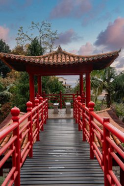 Kırmızı ahşap bir köprü Funchal, Madeira 'daki Monte Palace Tropical Garden' daki bir pagoda pavyonuna çıkar. Avuç içleri, kafes korkulukları, banklar ve gün batımı ışığı sakin bir ortam oluşturur..