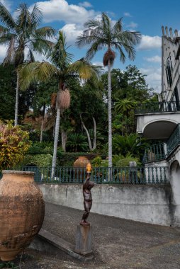 Funchal, Madeira, Portekiz 'deki Jardim Monte Sarayı, kemerli balkonları, yeşil korkulukları, yüksek palmiyeleri, yoğun eğreltiotları, bronz çocuk heykeli ve kilden bir vazosu olan bir malikane gösteriyor..
