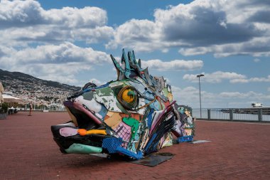Geri kazanılmış plastiklerden oluşan çok renkli bir balık heykeli Funchal deniz önü, teraslı yamaçlı evler ve marina görülebilir durumda. Yeşil enerji ECO sembolü.