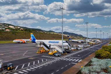 Madeira 'daki Cristiano Ronaldo Uluslararası Havalimanı Discover Airlines, Ryanair ve Easy Jet uçaklarının parçalı bulutlu gökyüzü altında mürettebat, yük arabaları ve merdiven kamyonlarıyla kapılarda uçtuğunu gösteriyor..