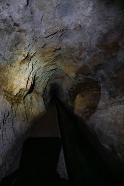 Portekiz 'in Madeira kentindeki dar bir kaya tünelinde kabataslak duvarlar ve sığ bir su kanalı görülüyor. Bir el feneri sert gölgeler, dünyevi tonlar ve birbirine yaklaşan çizgiler bırakır..