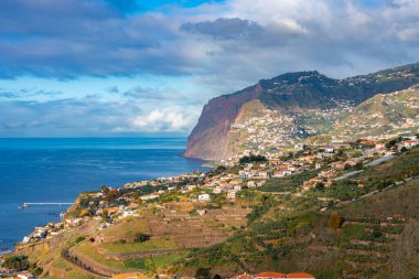 Portekiz 'in Madeira kentindeki Cabo Girao yakınlarında beyaz evleri olan teraslı yamaçlar Atlantik' e iniyor. Geç saatlerde güneş ışığı çatıları ve terasları ısıtır, bulutlar serin gölgeler bırakır..