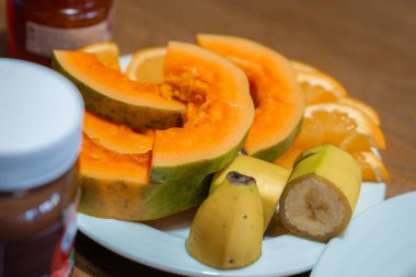 Funchal 'daki kahvaltı tabağını kapat. Papaya yeşil kabuklu, muz dilimli ve turuncu dilimli beyaz tabak, yumuşak iç mekan aydınlatmalı, sığ alan derinliğinde..