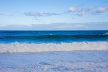 Portekiz, Madeira 'da köpüklü bir kıyı şeridine doğru yuvarlanan Atlantik dalgaları. Dağınık bulutlu soluk gökyüzü, katmanlı ufuk, yumuşak gün ışığı, dinamik dalgalar yakalanmış.