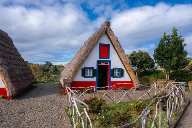 Portekiz 'in Madeira şehrinde bulunan geleneksel Santana Çerçeve Kulübesi kaldırım taşı yolları, yeşil kepenkleri ve parçalı bulutlu gökyüzünün altındaki uzak tepeleri olan temiz bir bahçede duruyor..