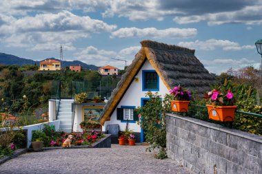 Geleneksel Santana sazdan çatı kulübesi beyaz duvarlar, mavi süslemeler ve yeşil panjurlar Funchal yakınlarındaki kaldırım taşından bir sokakta, bulutların altında çiçekler ve dağlar var..