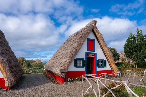 Beyaz duvarları ve kırmızı ve mavi süslemeleri olan üçgen sazdan bir kulübe Santana, Madeira, Portekiz 'de gün ışığı altında ve bulutlar altında kaldırım taşı bir yolun yanında duruyor..