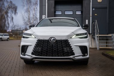 Beyaz Lexus RX 450 h artı ön yakın çekim Milli ızgara ızgara, L şekilli LED farlar, ve koyu panelli binanın yanındaki tuğla üzerine krom amblem kapalı kapılar.