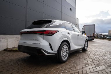 Beyaz Lexus RX 450 saat artı gri panelli binalar, tuğla döşemeler, park edilmiş kamyonlar, LED aydınlatma çubuğu ve alaşımlı tekerlekleri olan bir sanayi bölgesinde arka üç çeyrekten gösteriliyor..