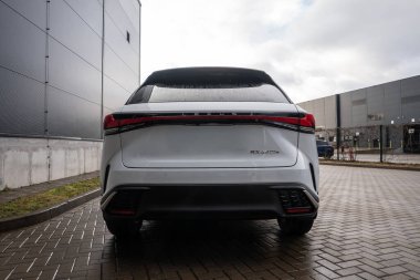 Beyaz Lexus RX 450 saat artı modern endüstriyel binalar arasında ıslak tuğlalı bir garaj yoluna park edilmiş. Su damlacıkları arka cama oturur, LED ışık çubuğu ve Lexus harfleri öne çıkar.