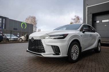 Beyaz bir Lexus RX RX 450 artı modern endüstriyel bir binanın yanındaki ıslak tuğla parkına park ediyor. Büyük bir ızgarası, LED farları, yükleme bölmesi kapısı ve yeşil dairesel bir tabelası var..