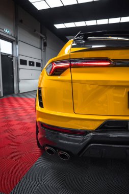 Lamborghini Urus 'un arka üç çeyreğinde kırmızı ve siyah kareli zeminde, LED arka lambaları, dört egzozu, yayıcıyı ve çatı spoiler' ını gösteren sarı bir Lamborghini Performante var..