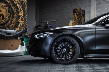 Üç çeyrek açıda siyah bir Mercedes Benz S Sınıfı görünüyor. Matet boyalı, parlak siyah tekerlekler ve sarı kalibreli, kapalı bir atölyede aletler ve duvar sanatıyla birlikte..