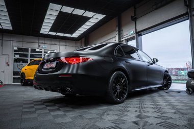 Sarı Lamborghini Urus Performante körfez kapılarının yanında dururken, siyah bir Mercedes Benz S sınıfı atölyede oturuyor. Checker döşeme, panel ışıkları ve bulutlu gökyüzü gösterisi.