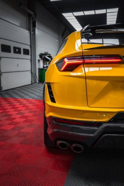 Dar üç çeyrek dikiz açısı gösteriyor ki, sarı Lamborghini Urus Performante kırmızı ve siyah kareli bir atölye zemininde park halinde LED arka lambası, dörtlü uçlu, dağıtıcı ve aero kanadı görünür..