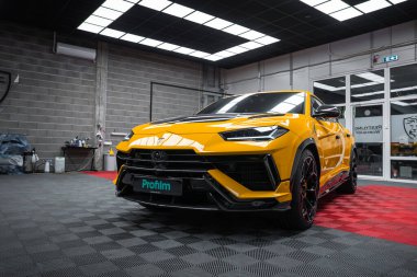 Sarı bir Lamborghini Urus Performante, kapalı bir Profilm stüdyosunda, LED ızgara ışıkları altında, parlak siyah aksanlı ve kareli zemin üzerinde kırmızı halka tekerlekleri ile park edilir..