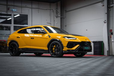 Parlak sarı Lamborghini Urus Performante gri kareli zeminde oturuyor kırmızı şeritli, siyah ayırıcı, karbon aksanlı ve yumuşak ışık altında büyük siyah tekerlekler.