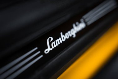 Sığ bir derinlik çekimi, parlak siyah bir kapı eşiğinde parlak bir Lamborghini betiği gösterir, canlı sarı köşegen aksanı ve dar odak içinde karamsar düşük ışıklandırma.