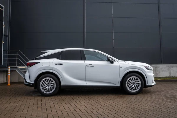 Beyaz Lexus RX 450h artı koyu metal kaplı endüstriyel bina ve merdivenlerin yanındaki tuğla parka park edilmiş F spor rozetleri, alaşımlı tekerlekler, keskin çizgiler ve bulutlu ışıkta melez tasarımı gösteriliyor..