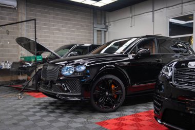 Parlak siyah Bentley Bentayga Speed bir tamircide oturuyor, elmas ızgara ızgara, oval far, siyah beş tekerlekli, turuncu kalibre, asansör ve aletler görünür..