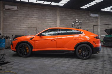 Orange Lamborghini Urus SE bir atölyede, profil görünümünde, altıgen kemerler, siyah çoklu jantlar, turuncu kalibreler, beton duvarlar, şerit aydınlatma, mevcut diğer arabalar.