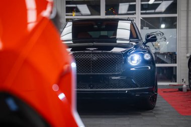 Parlak siyah Bentley Bentayga Speed kapalı bir garajda kameraya, matris ızgarasına ve tek LED lambaya, ön tarafta turuncu araba bulanıklığına, ışıkların altında fayans ve kırmızı paspaslara bakıyor..