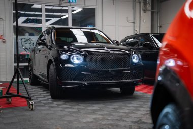 Parlak siyah Bentley Bentayga Speed kameraya kapalı alanda, ızgarada ve oval LED farlarda parlıyor. Atölye ışıklandırması, lastik döşeme ve teçhizatlar sahneyi hazırladı..