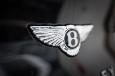 Yakından bakınca, parlak siyah Bentayga Speed 'in kanatlı krom Bentley B amblemi görünüyor. Yumuşak bulanıklık, sergi odası tarzı ışıklandırma altında yontulmuş tüyleri ve yansımaları izole eder.