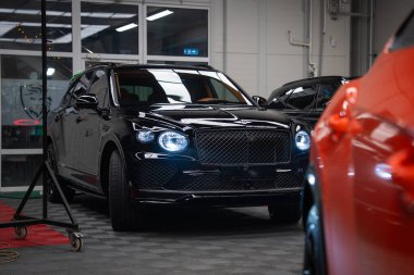 Bentley Bentayga Speed, matris ızgarası ve dairesel LED farları parlayan, turuncu performans arabası kısmen görünür, serin dağınık ışıklandırmalı bir bölmede oturuyor..