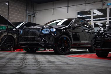 Parlak siyah Bentley Bentayga Speed kareli zeminde oturuyor, matris ızgara ve oval LED farlar görünür, siyah tekerlekler, kırmızı kalibreler, aletler ve duvarlar garaj ayarlarını gösteriyor.