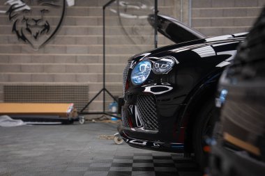 Siyah Bentley Bentayga Speed, kapalı bir atölyede, tuğla duvarlı, aslan başlı amblemli ve kareli zeminde, hatta serin ışıklandırma altında yükseltilmiş kaput ve LED farları gösteriyor..