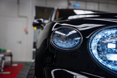 Kapalı alanda siyah Bentley Bentayga Speed 'in sol ön far kümesini gösteriyor. Çift dairesel LED lambaları ve yukarıdan yansıyan kristal ışıkları var..