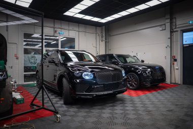 İki siyah Bentley Bentayga Speed SUV kapalı bir bölmede park halinde. Yukarıdaki floresan paneller boyaya yansıyor, oval farlar ve matris ızgarası görünür..
