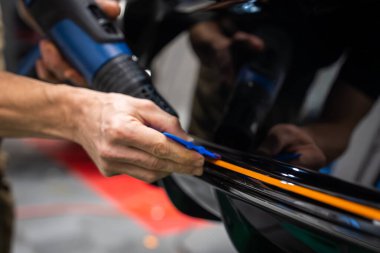 Otomobil servis hangarında ellerinizi kapatın. Parlak siyah bir panele turuncu bir vinil şerit uygulayın. Isı tabancası ve parlak ışık altında mavi cam sileceği kullanın..