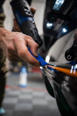 Bir araba teknisyeni kapalı alanda parlak siyah bir araca mavi bir cam sileceği ve ısı tabancası kullanıyor. Eller portakal şeridine odaklanır, kareli zemin ve parlak iş ışıkları görünür..