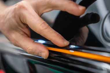 Bir işçi atölyede parlak siyah bir araba paneline turuncu çizgili bir çizgi çiziyor. Dar çerçeve, sığ alan derinliği ve yumuşak ışıklandırma dikkatli el basıncını gösteriyor.