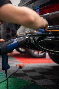 Bir teknisyen parlak siyah bir araba tamponuna ısı tabancası ve cam sileceği kullanarak vinil ambalaj uygular. Kırmızı arka lamba, krom kaplama, kareli zemin ve kırmızı paspaslar görünür..