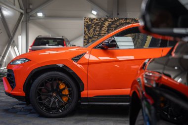 Orange Lamborghini Urus SE iyi aydınlatılmış bir atölyede oturuyor. Ön tarafta siyah çoklu konuşma tekerlekleri, turuncu fren kalibreleri, keskin çamurluk menfezi ve parlak yansımalar var..