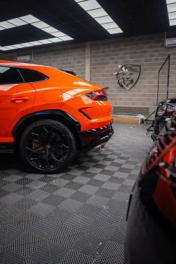 Turuncu Lamborghini Urus SE, aslan logosu, kareli döşeme ve panel ışıkları olan bir atölyede tekerlek, turuncu kalibre ve LED stop lambası gösteriyor..