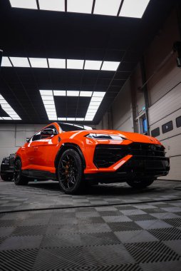 Modern bir atölyede turuncu Lamborghini Urus SE 'nin düşük açılı görüntüsü, LED çalışan ışıklar ve altıgen ızgara ileri, kauçuk şebeke zemin ve tavan panelleri vücut üzerinde yansıyor.
