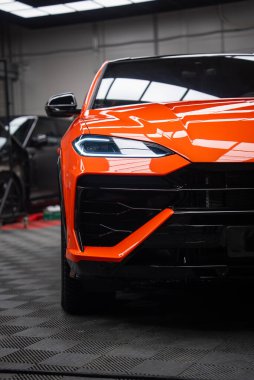 Kapalı mekandaki bir atölyede turuncu Lamborghini Urus SE 'yi gösteriyor. Açısal LED far, oyulmuş kaput, siyah ızgara ve parlak ışıkların altında turuncu aero bıçağı var..