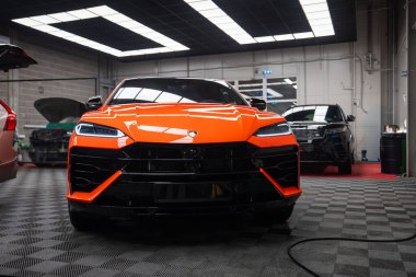 Canlı turuncu bir Lamborghini Urus SE iyi aydınlatılmış bir atölyede duruyor. Açısal fasya, boğa arması ve siyah ızgara göze çarpıyor. Yakındaki SUV ve açık kaputlu araba servis işi öneriyor.