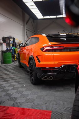 Turuncu bir Lamborghini Urus SE 'nin dörtte üçlük dikiz görüntüsü kapalı park halindeki arka lambaları, dört egzoz uçlarını, siyah difüzörü ve parlak siyah tekerlekleri gösteriyor..