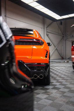 Bir atölyede canlı turuncu Lamborghini Urus SE arka çeyreği, dörtlü egzozlar, keskin stop lambaları, parlak siyah difüzyon, kareli lastik zemin, siyah ve kırmızı diğer arabalar